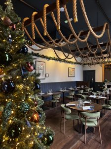 A decorated restaurant interior with a large Christmas tree, festive garlands, and overhead rope lighting